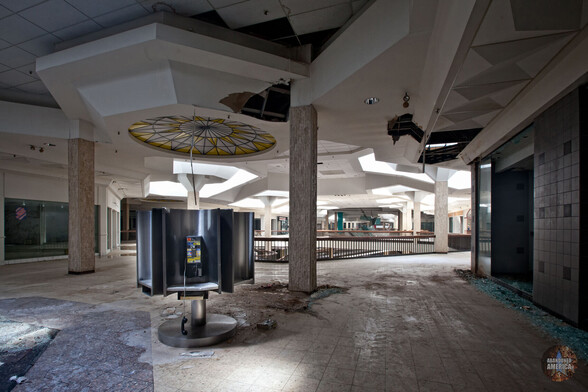 A darkened stretch of the second floor of an abandoned mall. A payphone kiosk is in the foreground, one of the receivers dangling on its cord. In the background there is a disorienting mixture of ramps and skylights, all at odd angles