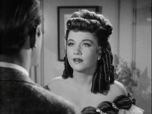 A woman wearing an 1800s ringletted hairstyle and a low-cut dress is standing in the dressing room of a theater talking to a man (he is facing her with his back to the camera)