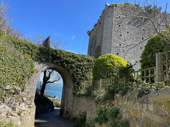 Rufus Castle on the east coast of Portland. An old, square rugged granite citadel that real men dream about after a hard day working on spreadsheets. The sky is blue behind. The sea visible through an ivy-covered archway. 