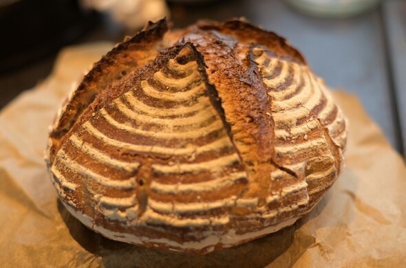 A loaf of homemade sourdough bread fresh from the oven. It has pointy ears when the crust split along the cuts at the top and is dark brown, with rings of flour.