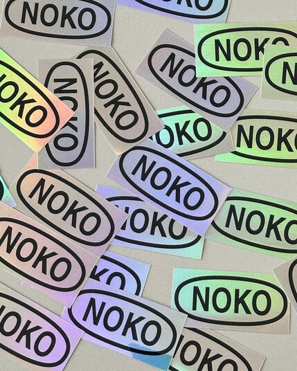 lots of silvery holographic stickers containing an oval shape with "noko" in it, on a light surface.