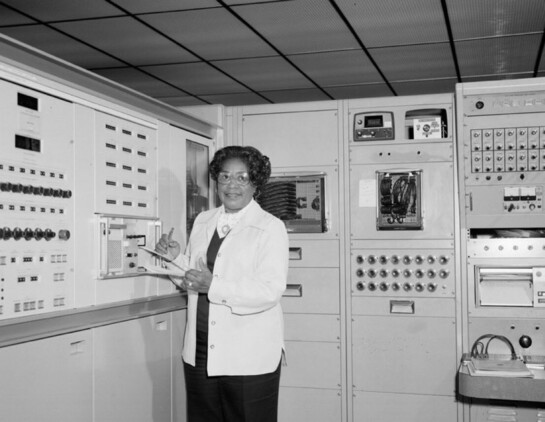 A black and white photo of Mary Jackson standing in front of a bank of floor-to-ceiling computers in the 1960s. She has short curly hair and is wearing glasses and a white lab coat. Jackson appears to be pulling a wire from a panel on one of the computers.