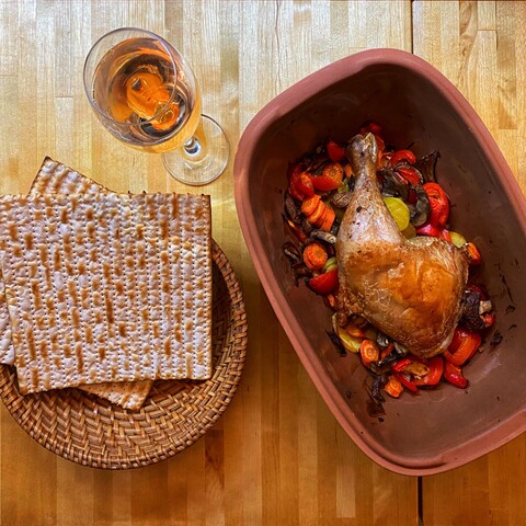 Top-down image of a wooden table with food and drink: a roasted chicken leg with vegetables in a clay pot; matzot in a basked; and a glass of rosé.