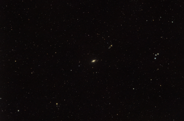 This is a photograph of the Sombrero Galaxy (M 104), which appears as a luminous disk bisected by a thin, dark line. Surrounding the galaxy are several colorful and bright stars.