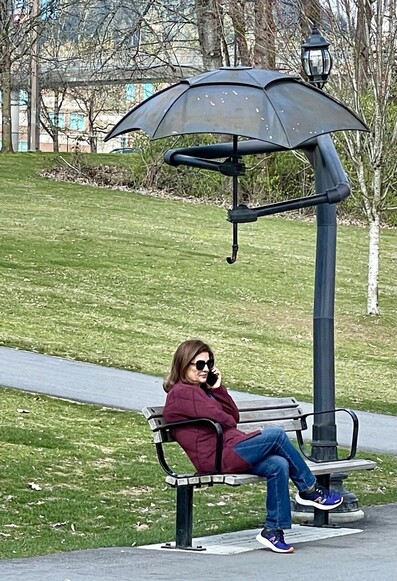 Woman on rainy day sitting on a park bench on phone under a whimsical modern sculpture of a person holding an umbrella over her. 