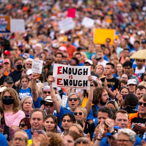 Photograph of hundreds protesting. One sign stand out in the middle which says enough is enough.