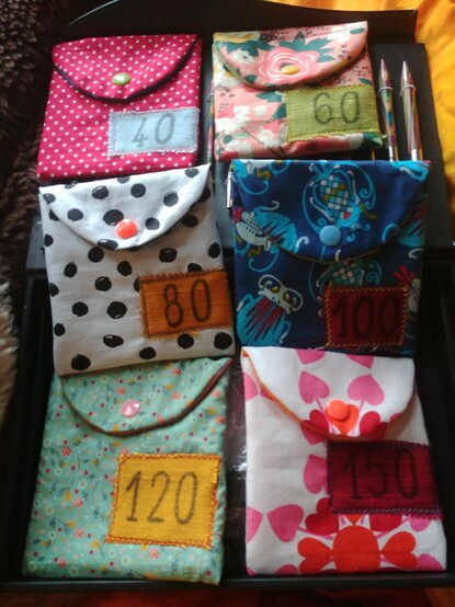 Sechs bunte, genähte Täschchen mit Drucknopf, jeweils unten rechts eine aufgestempelte Zahl (40, 60, 80, 100, 120, 150), die in zwei Reihen von je drei Stück nebeneinander liegen.