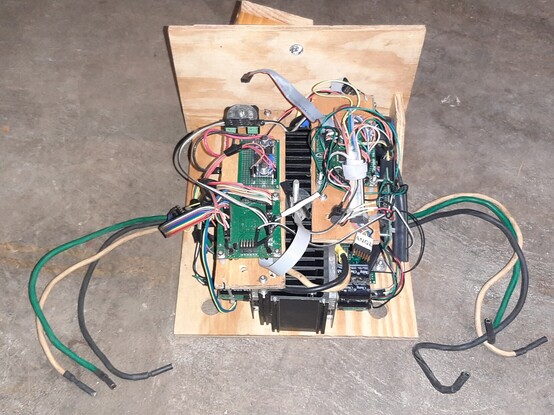 Two scratch built motor controllers mounted to a heat sink mounted to a plywood box enclosure. Out of the kart and sitting on the concrete floor. Three phase wires hang out from each side. Lots of ribbon cables and colorful dupont wires in a messy configuration with velcro ties.
