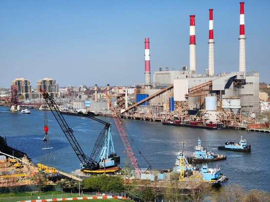 Three tugboats attend to two barges each carrying cranes. The larger barge mounted crane is lifting a third crane onto land. Across the water a large fossil fuel power plant looms, with four striped smokestacks pointing skyward.