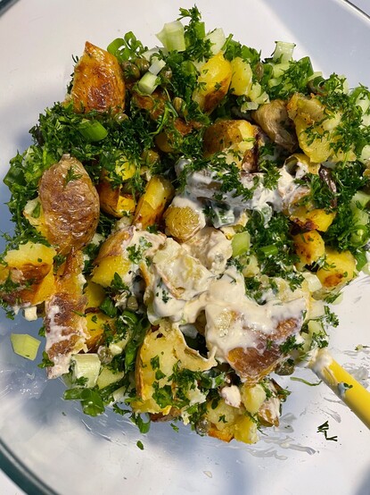 Combining the ingredients for the roasted Yukon gold potato salad in a clear glass bowl. There are roasted potatoes, minced parsley, dill, green onions, and the sour cream dressing as it is being mixed into the salad.