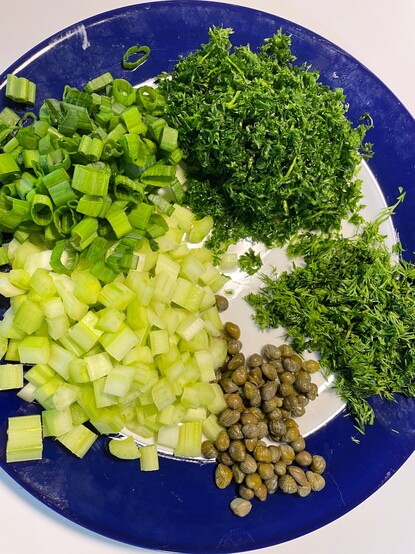 The greenery to be added to the potato salad, sitting on a blue plate. Top left and clockwise: green onions, parsley, dill, capers, and celery, all chopped up and ready to go. 