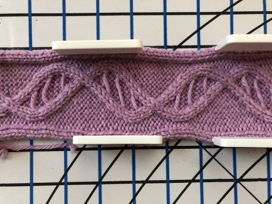 Close up photograph of a section of pink cabled knitting resembling a DNA double helix. It is pinned down to a white piece of foam used to block the knit fabric so that it will lie flat. The foam mat has parallel lines and concentric circles to aid in blocking projects to the correct dimensions and shape.