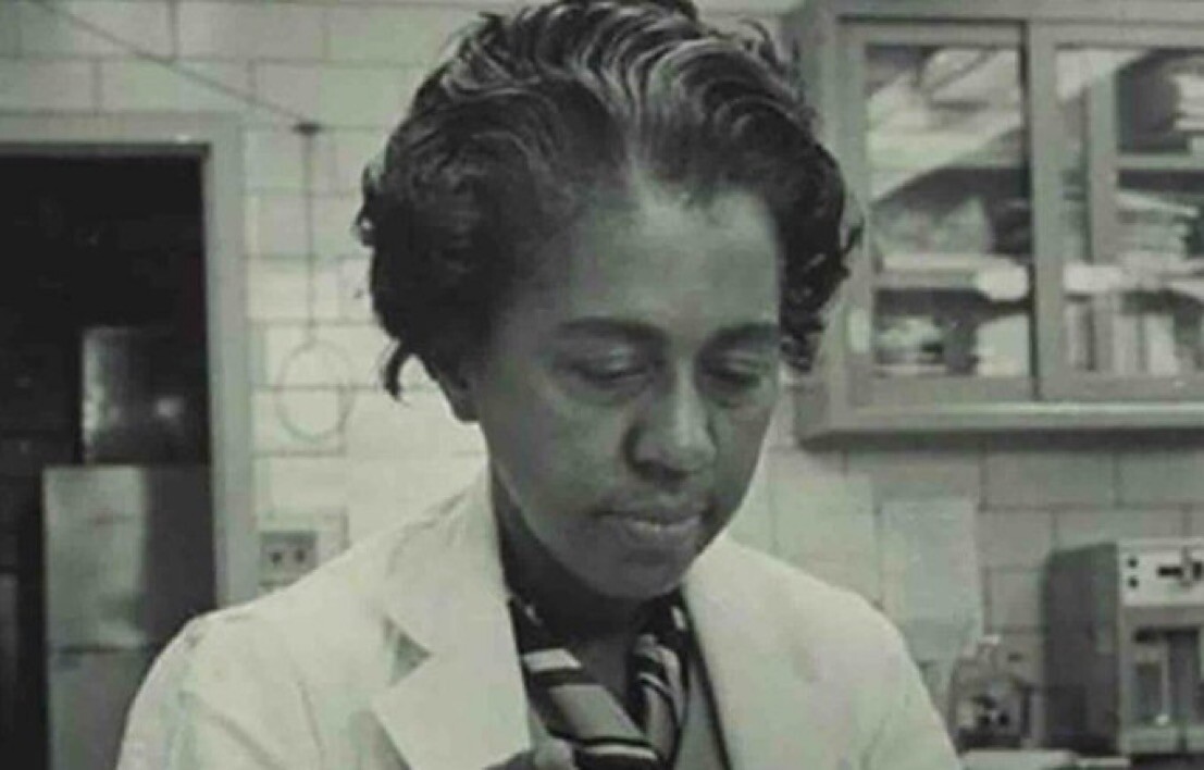 A black and white photo of Marie Maynard Daly. It shows her from the shoulders up, wearing a white lab coat with a striped top underneath. She is in a lab; there are white tile walls, glass cabinets, and some lab equipment in the background. She is looking slightly downward, probably at something she is holding in her hands.