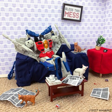 The transformer figure Starscream sitting on a messy sofa, in messy room, with cats and papers around them. And a wonky picture which says ‘ Bless this mess’