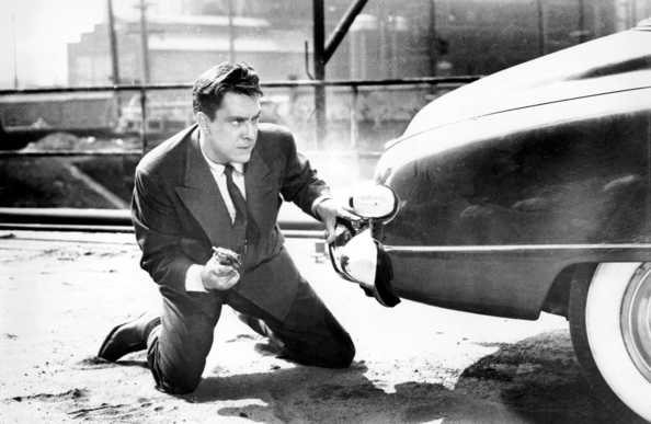 A man wearing a 40s style suit and tie is on his knees behind a car with one hand on the bumper and the other holding a gun