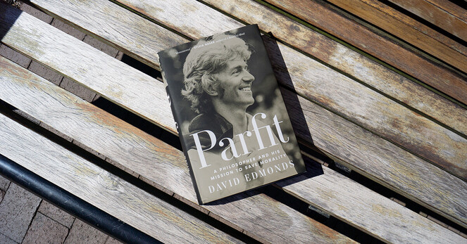 Parfit by Dave Edmonds lays on a wooden slatted bench. The cover is a black and white photo of Derek Parfit smiling and facing right with his jacket draped over his left shoulder. 