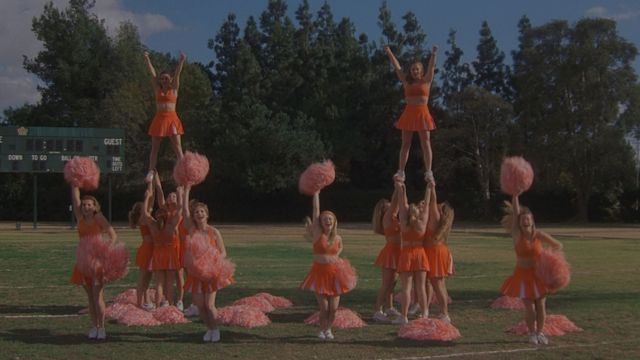 A high school cheerleading team is practicing a cheer routine on a football field. They are wearing orange cheerleading uniforms and have pompoms. Four girls are cheering in the front row. Behind them are two groups of four cheerleaders each who are holding another cheerleader up in the air.