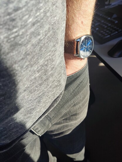 Foto van een hand in een broekzak, met aan de pols een horloge van het merk Seiko met een blauwe wijzerplaat die lichte en donkere tinten blauw gecombineerd weergeeft door de manier waarop de zon op het horloge invalt. De bruine kleur van het lederen bandje is ook nog net zichtbaar.