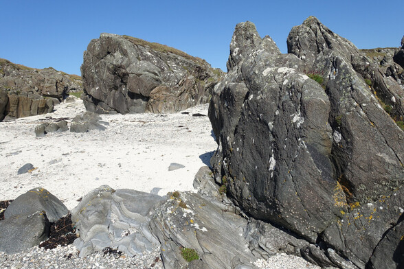 Gnarly outcrops of grey and black rocks surround a beach of white sand, under a blue sky. The rocks in the left foreground show banding of paler and darker grey and are metamorphosed sedimentary rocks. The darker rocks to the right, and in the centre background are garnet amphibolite, originally a basaltic sill or dyke cutting the sedimtnary rocks, before they were all metamorphosed. In the dark amphibolite near the contact with the banded grey rock, pinkish spots of garnet can be seen: they are much bigger near the contact than inside the amphibolite. This metamorphism probably occurred around 450 million years ago.