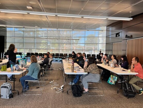 A group of instructors sit at three long tables with their laptops as a colleague shows them how to use LibreTexts to create, remix, and manage their OER projects.