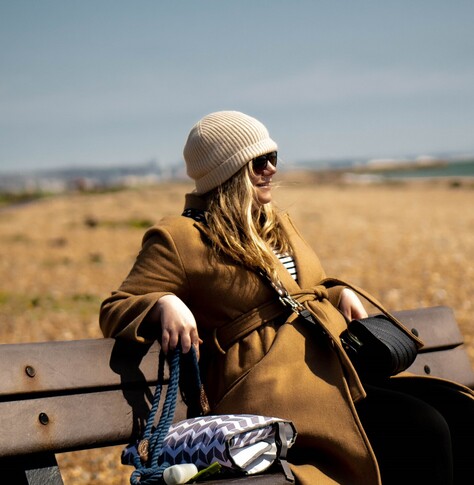 Sideways 3/4 length image of my daughter sitting on a bench at the beach at Shoreham, wearing a brown coat and a beige beanie. 