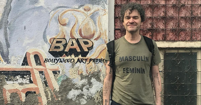 A picture of Mark Cousins in from of a wall with a mural saying BAP Bollywood Art Project. He wears a shirt that says 'Masulin Feminin' referring to the Godard film of that name.