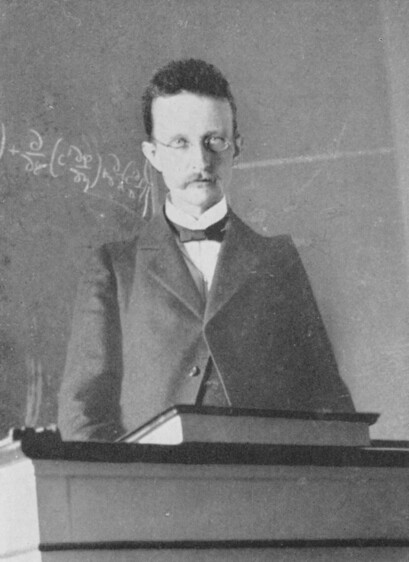 A young Max Planck standing at a lectern. He is in his early to mid twenties, wearing a dark jacket that is buttoned high, a white shirt, and a black tie similar to a bow tie. Planck has thin-rimmed glasses and a substantial mustache.There is an equation written on the blackboard in the background.
