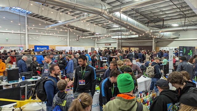 Event room with tables of 3D printed projects. Hundreds of people walking through talking.
