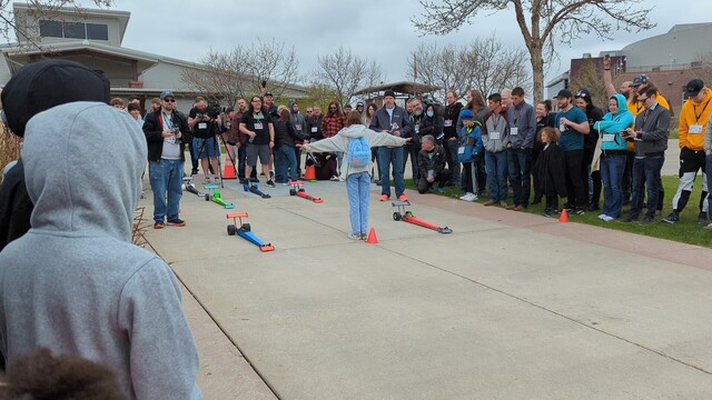 Crowd around a sidewalk with 3D printed drag race cars lined up to race. 