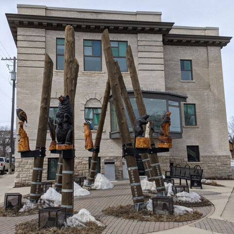 Sculpture in front of a brick building of seven large wooden poles in a circle with wooden animals sitting half-way up the poles 