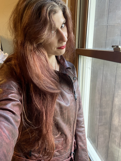 A girl with long, light red hair wearing red lipstick and a brown leather jacket, standing next to a window 