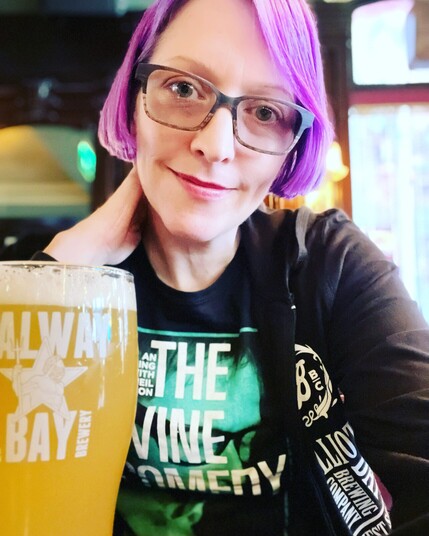 Your author, a pale white lady with purple hair and glasses, in at the bar in The Oak, in a black Divine Comedy t-shirt, with a Galway Bay pint in front of her.
