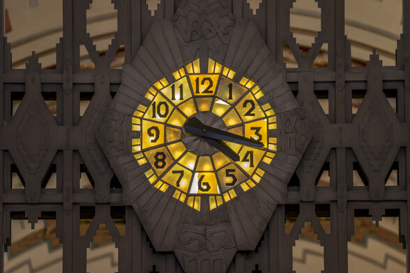 A photograph of the hand dial clock situated above the entrance gates to the retail promenade area of the Guardian Building in Downtown Detroit. The clock is integrated into a wrought iron gate that is a dark brown color. The face of the clock is backlight with a yellow-ish light. There are various etchings surrounding the clock face that radiate outward. The typeface for the numbers on the clock is highly distinctive and similar to other typefaces found throughout the building. The typeface features sharp path deviations where the shapes would normally be smooth curves.