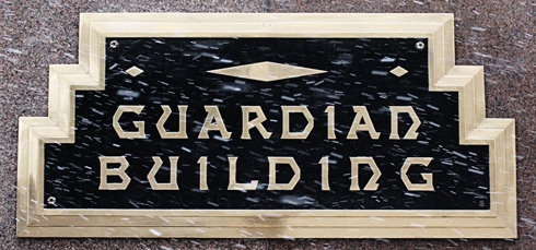 A plaque mounted outside of the Guardian Building in Downtown Detroit. The plague shape has a "staircase" pattern that is mirrored on both sides. Each staircase has three landings - which makes the width of the plaque on the bottom larger than the width of the top. The plaque features a think gold trim along its outer edges. Otherwise, the plaque is painted solid black with raised "Guardian Building" letter that is the same color as the outer gold trim. The typeface is distinctive and features sharp path deviations where the shapes would normally be smooth curves.
