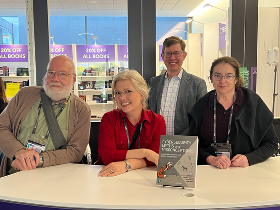 Gene Spafford, Pattie Spafford, Josiah Dykstra, and Leigh Metcalf standing behind a counter with a copy of their book on display.
