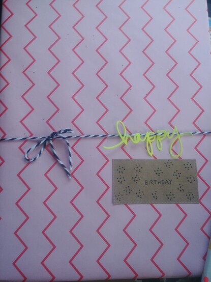 Verpacktes Geschenk.
Rosa Papier mit pinken Zacken.
Schwarzweißes Garn zur Schleife gebunden, an dem in neongelb das Wort happy hängt.
Ein rechteckiges Stück Kraftpapier klebt unter dem Wort happy auf das das Wort Birthday und Pünktchen gestempelt wurden.