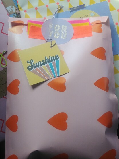 Verpacktes Geschenk.
Rosa Papiertüte mit orangenen Herzen, zugeklebt it Textilband in Neonfarben.
Ein runder Aufkleber mit der Aufschrift For You und ein Geschenkanhänger mit dem Aufdruck Sunshine dienen zur Deko.