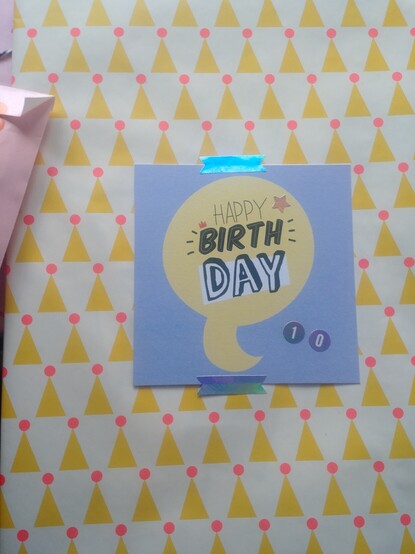 Verpacktes Geschenk mit einem Papier, das ein Geburtstagshütchen ( in gelb mit neonorangenem Bommel) Alloverdruck zeigt.
Darauf klebt ein bedrucktes Papier mit der Aufschrift Happy Birthday.