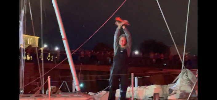 A smiling Kirsten Neuschäfer waves at the crowds on the quay of Les Sables d’Olonne, France, from the foredeck of her sailing boat ‘Minnehaha’. It’s night, and she has a red plastic horn in her hand.