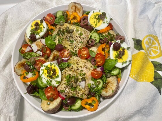 Overhead view of a mock Salad Niçoise, using mahi mahi instead of tuna. There are hard-boiled egg halves topped with anchovy bits, roasted red potatoes, kalamata olives, baby bell pepper slices, cherry tomatoes, fresh basil. Sadly, I had n green beans. 