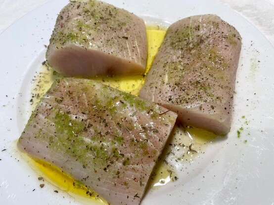 Mahi mahi marinating on a white plate in garlic-infused olive oil, garlic-scape powder, salt & pepper.