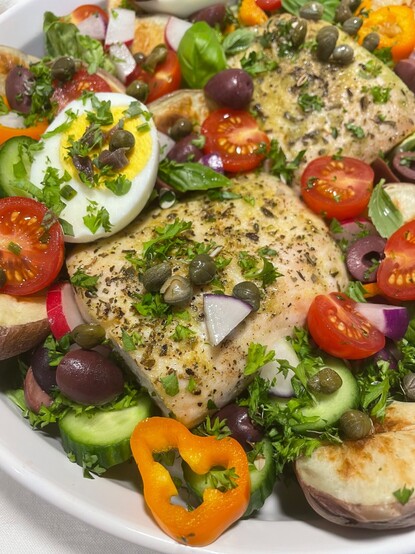 Close-up of the mahi mahi in the Salad Niçoise, with capers, cherry tomatoes, herbs, cucumbers, and radish bits. There is a hard-boiled egg half, topped with capers and anchovies. 