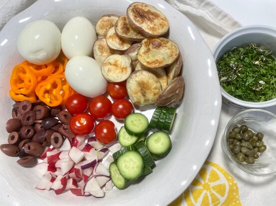 Mise en place. The available salad ingredients on a white plate: hard-boiled eggs, roasted red new potato halves, cherry tomatoes, cucumbers, radishes, kalamata olives, and orange baby bell peppers. To the right are little bowls of chopped fresh herbs and capers.
