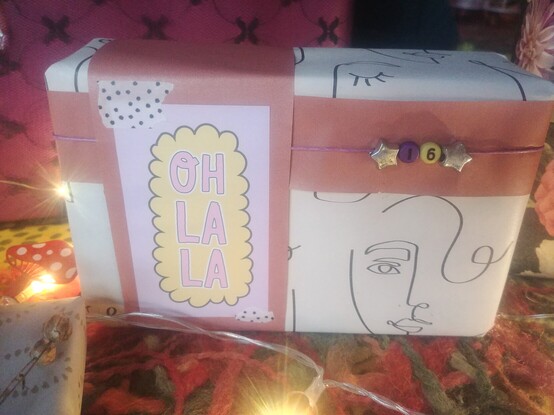 Verpacktes Geschenk mit Gesichtern in Online-Art.
Eine Karte mit der Aufschrift Oh la la ist mit gepunktetem Washitape aufgeklebt.
Ein Band mit Sternenperlen und einer 16 aus Perlen ist um das Geschenk gebunden.