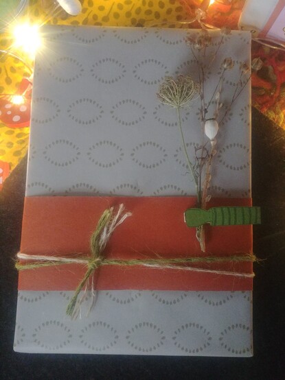 Ein in grau-grüne gemustertes Papier verpacktes Geschenk.
Ein terracottafarbener Papierstreifen ist im unteren Drittel um das Päckchen geklebt, drumherum ein doppeltes Juteband in grün und Naturfarbe.
Eine Wäscheklammer mit aufgeklebtem, gestempeltem Arm ist aufgeklebt und hält ein Sträusschen Trockenblumen.