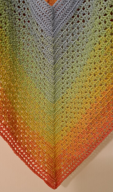 Ein Ausschnitt aus einem gehäkelten Poncho - oben sieht man gerade noch den Halsausschnitt, unten den Saum. Oben ist das Muster dicht, in der Mitte sind mehr Löcher und unten sehr viele. Die Farben gehen ineinander über: oben hellblau, dann hellgrün, dann gelb, unten hellrot.