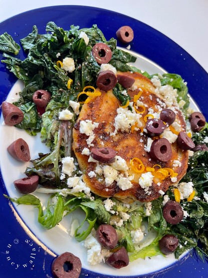 A blue-rimmed plate with a savory pancake on a bed of mixed, sautéed greens. Slices of kalamata olives, lemon zest and feta crumbles top it all off. 