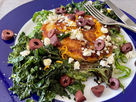 Another picture of the blue-rimmed plate with a savory pancake sitting on a bed of sautéed, mixed greens. The salad is topped with kalamata olive slices, lemon zest, and feta crumbles.