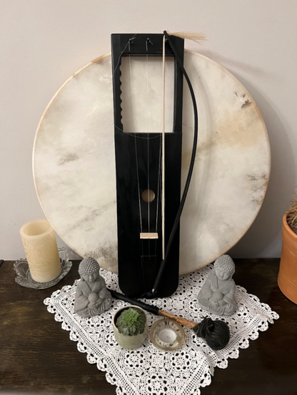 A sideboard with a large shaman drum and a black Talharpa in front of the drum.
The bow is hanging from the edge of the instrument

