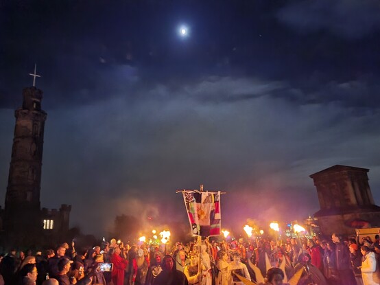 Part of the Beltane Fire Festival heads towards the camera, flaming torches and a banner held aloft the costumed and made up cast of fantastic characters are flanked by a crowd left and right, behind and above them a large moon lighting clouds in the sky, top left the Nelson monument tower sticks up and middle right an old stone building of the city observatory stares impassively down